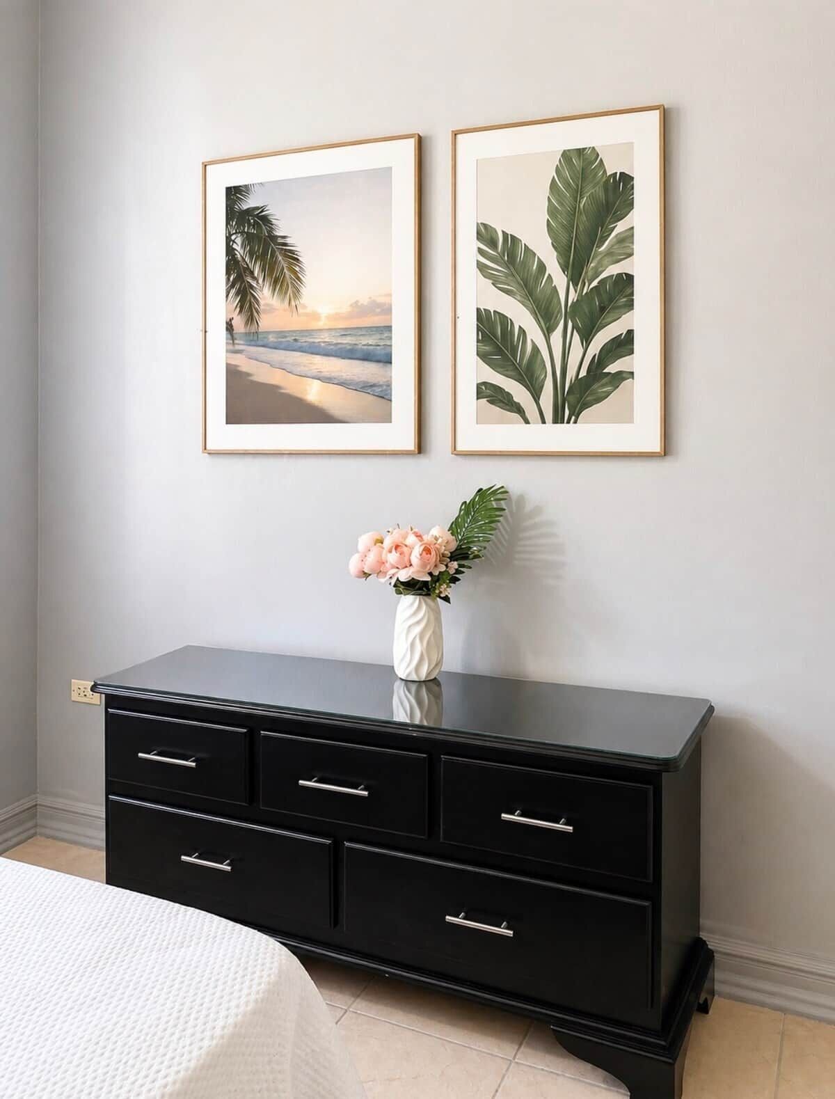 Une chambre à coucher avec une commode noire, deux tableaux encadrés accrochés au mur et un vase rempli de fleurs posé sur la commode.