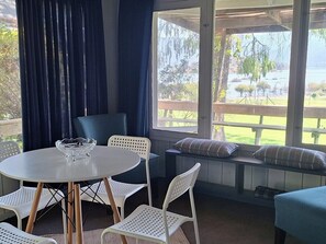 Un espacio de estar al aire libre cubierto, con una mesa y sillas, un banco con cojines, y vistas a un lago y follaje.