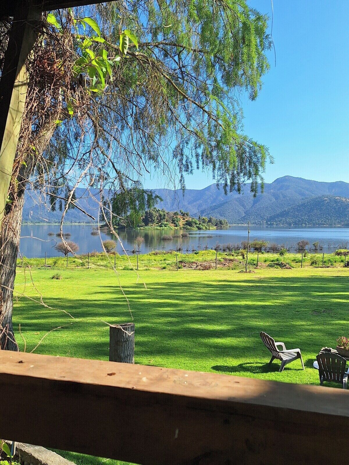 A grassy outdoor area with a wooden deck and two chairs, overlooking a calm lake and distant mountains.