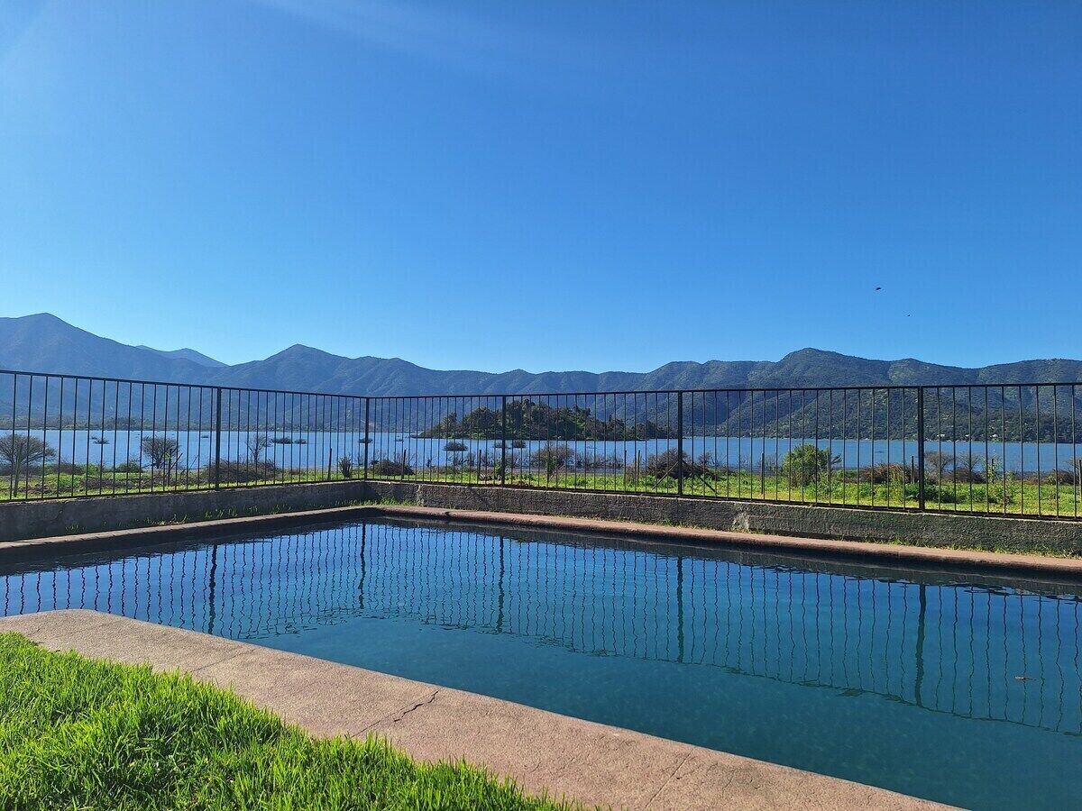 A rectangular swimming pool with a metal fence, surrounded by grass and a view of mountains and a lake.