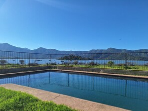 A rectangular swimming pool with a metal fence, surrounded by grass and a view of mountains and a lake.