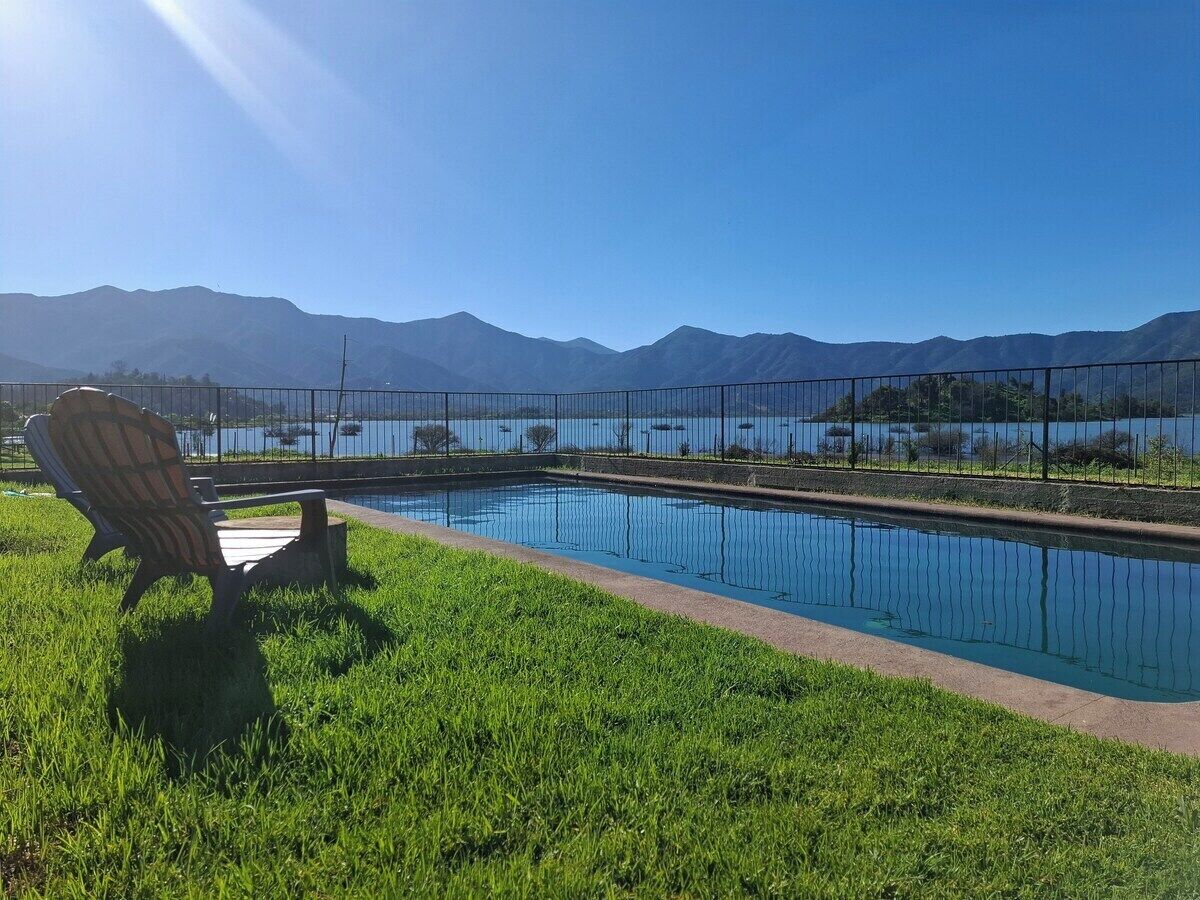 Una piscina con una silla y una valla, rodeada de césped y montañas al fondo.