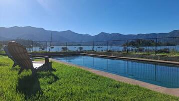 Una piscina con una silla y una valla, rodeada de césped y montañas al fondo.