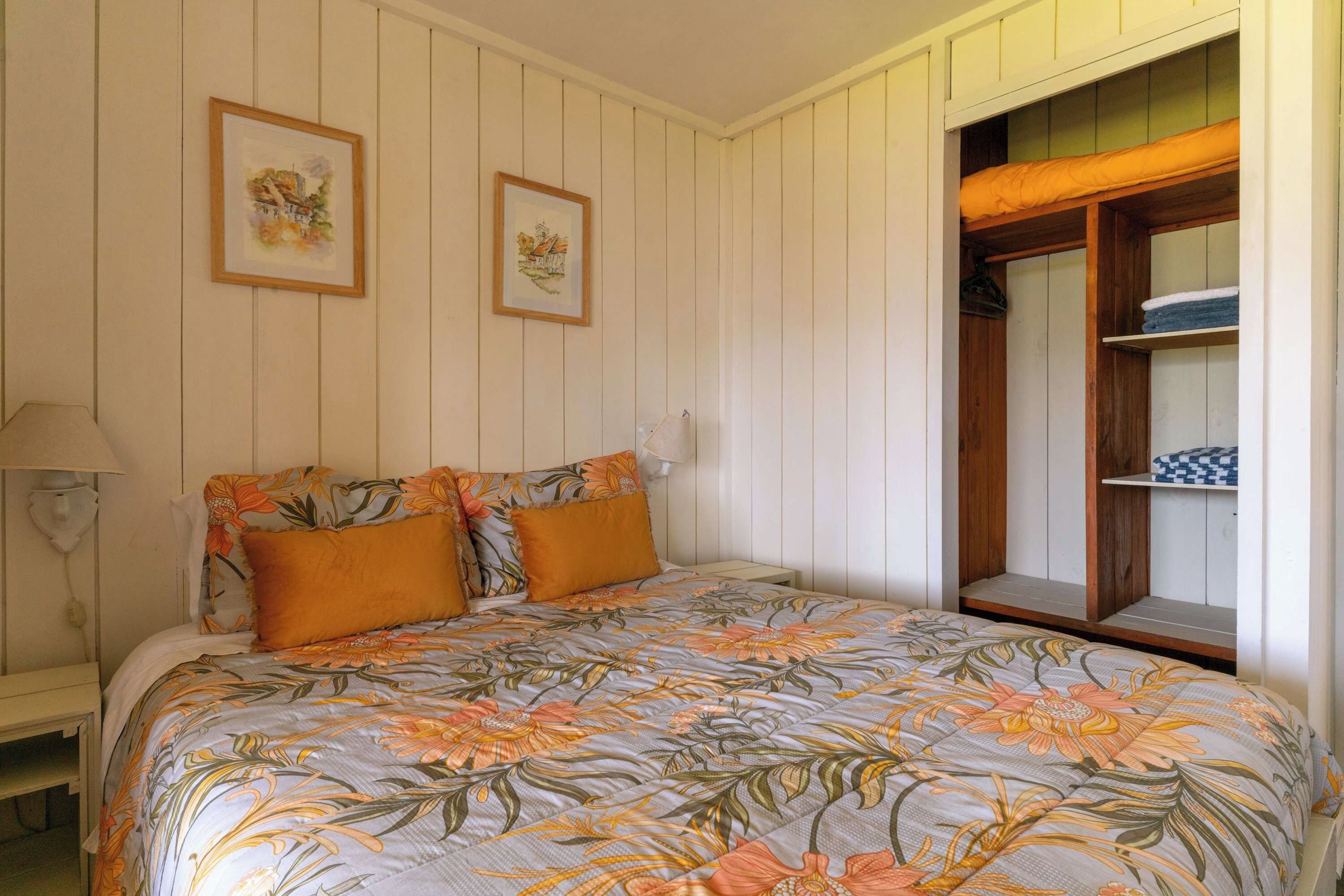 Une chambre à coucher avec un lit, deux tableaux encadrés au mur, une table de chevet et une armoire en bois avec une fenêtre.