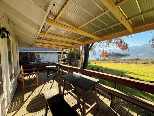 Una terraza techada al aire libre con mobiliario de madera, vista a un lago y un paisaje montañoso al fondo.
