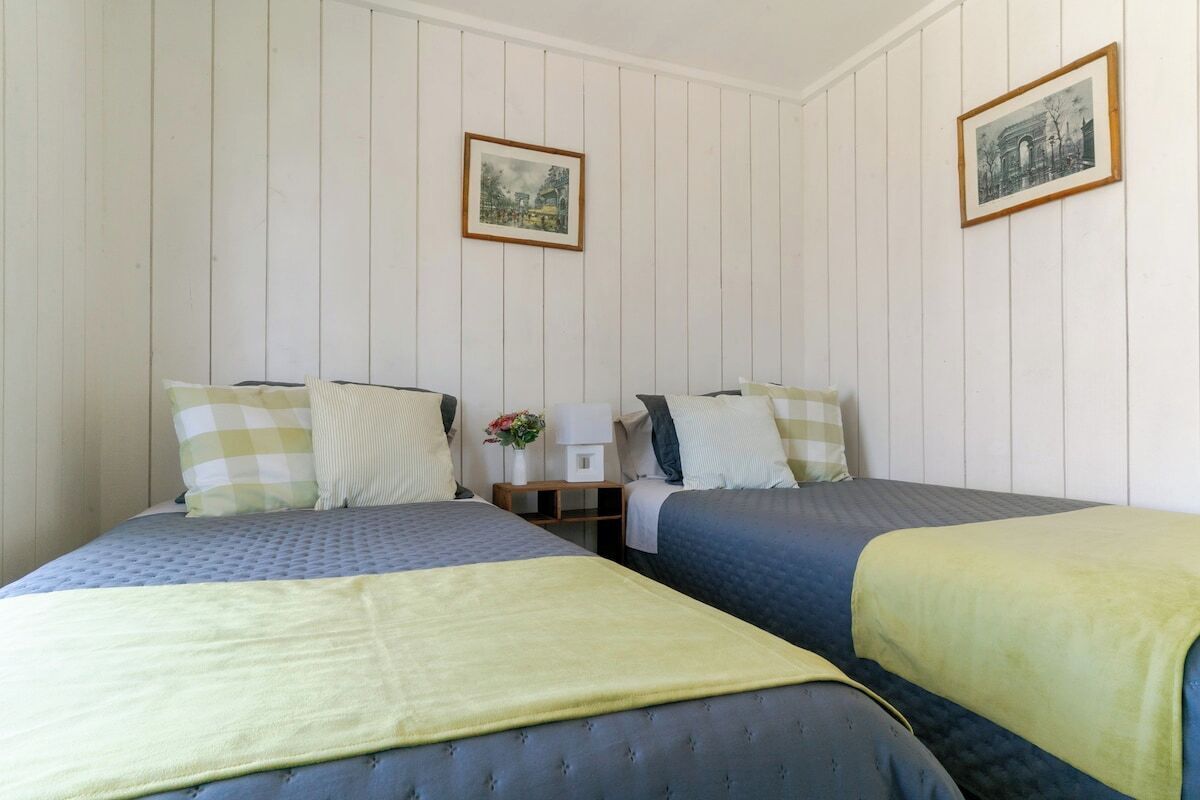 Una habitación con dos camas individuales, una mesita con un jarrón de flores y dos cuadros en la pared.