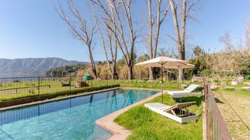 Piscina con sillas de descanso y una sombrilla, rodeada de árboles y con vistas a montañas.