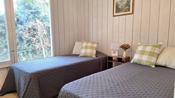 Une chambre avec deux lits, une fenêtre donnant sur des arbres, un tableau encadré au mur et une petite table sur laquelle est posé un vase de fleurs.