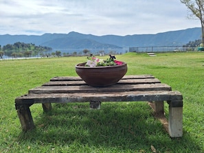 Um banco de madeira com um vaso de flores no centro, situado em um campo gramado com um lago e montanhas ao fundo.