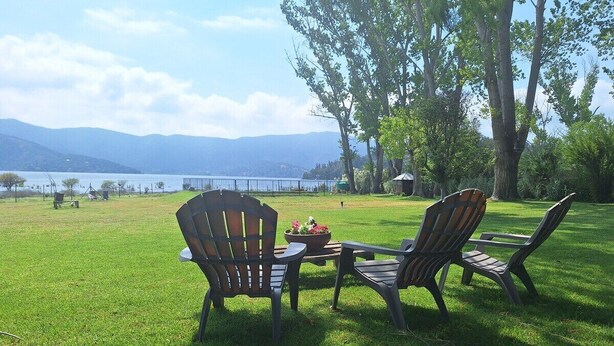 A grassy outdoor area with two Adirondack chairs and a table, overlooking a body of water and distant mountains. - Lakefront Cabin: Hot Tub, Pets OK, Near Santiago (Maipo)