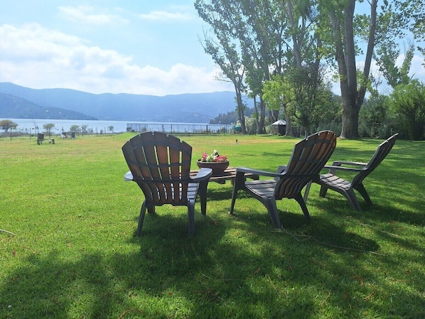 Área gramada ao ar livre com duas cadeiras Adirondack e uma mesa, com vista para um corpo d'água e montanhas ao longe.