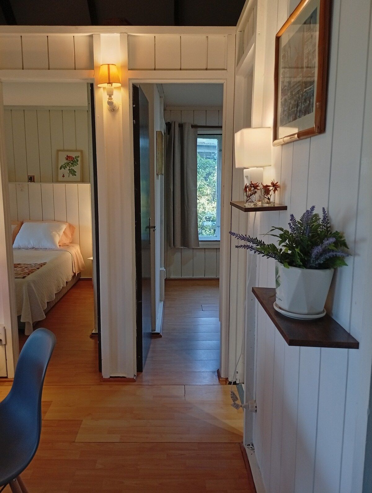 Una habitación con cama, una silla, un escritorio, un estante con plantas y una puerta que da a otra estancia.