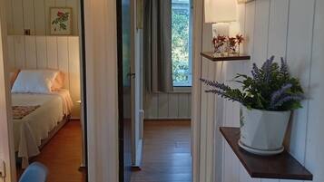 Una habitación con cama, una silla, un escritorio, un estante con plantas y una puerta que da a otra estancia.