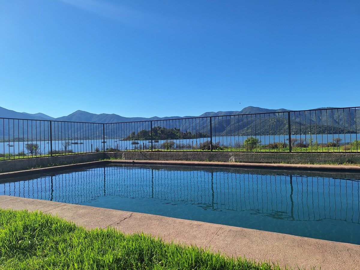 Una piscina rectangular con superficie de agua azul cristalina, rodeada por una valla metálica, y vistas a montañas y un lago al fondo.