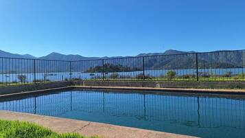 Una piscina rectangular con superficie de agua azul cristalina, rodeada por una valla metálica, y vistas a montañas y un lago al fondo.