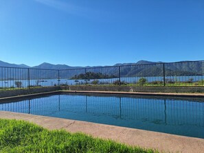 Piscina retangular com superfície de água azul cristalina, cercada por uma grade de metal, e vista para montanhas e um lago ao fundo.