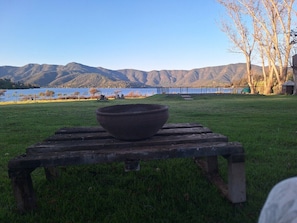 A wooden table with a bowl on it, set on a grassy area with a view of mountains and a body of water in the background.