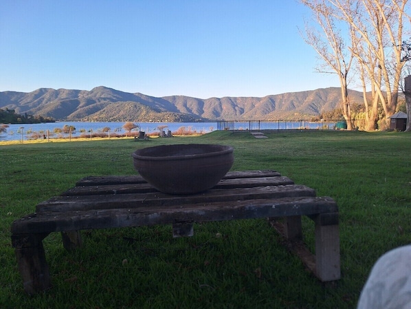 A wooden table with a bowl on it, set on a grassy area with a view of mountains and a body of water in the background.