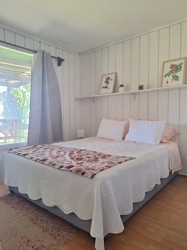 Une chambre à coucher avec un lit, une fenêtre avec des rideaux, une étagère fixée au mur avec des tableaux, et un tapis à motifs.