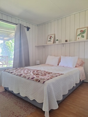 A bedroom with a bed, a window with curtains, a wall-mounted shelf with pictures, and a patterned rug.
