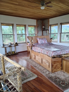 A wooden bed with storage drawers, a ceiling fan, and large windows with a view of trees.