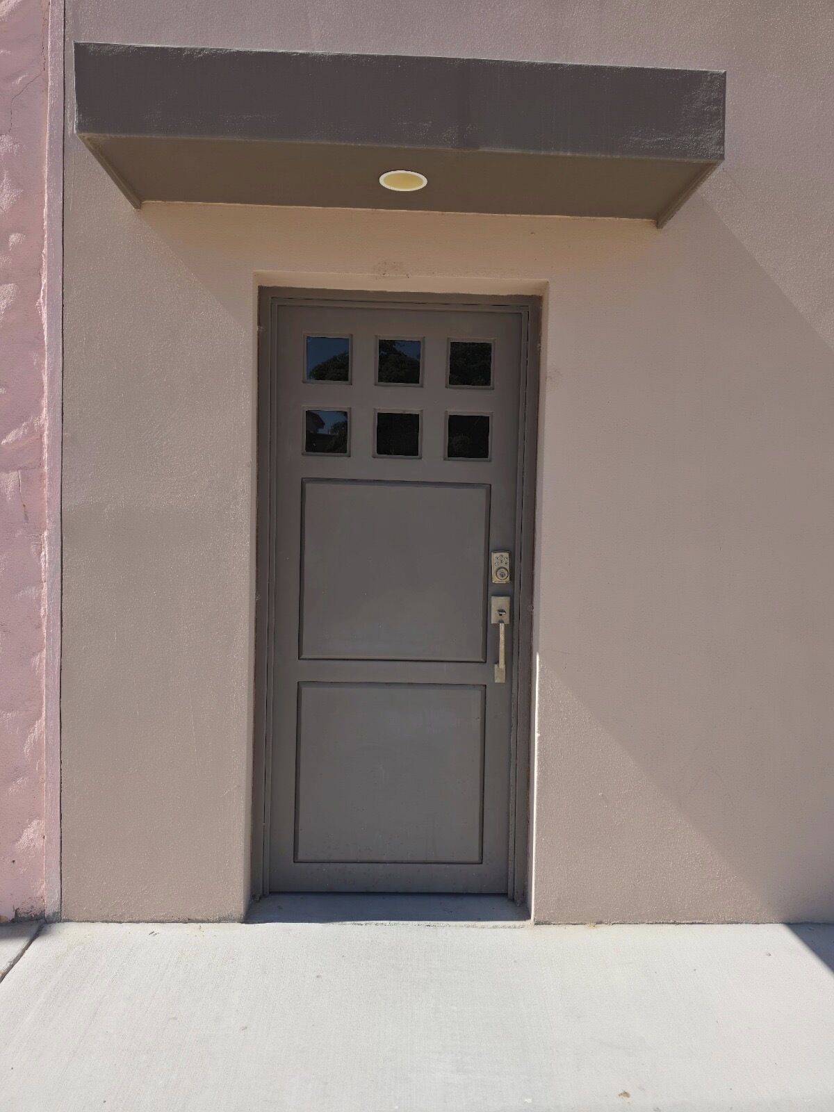 A grey door with a keyhole and a handle, set in a building with a concrete base and a pinkish wall.
