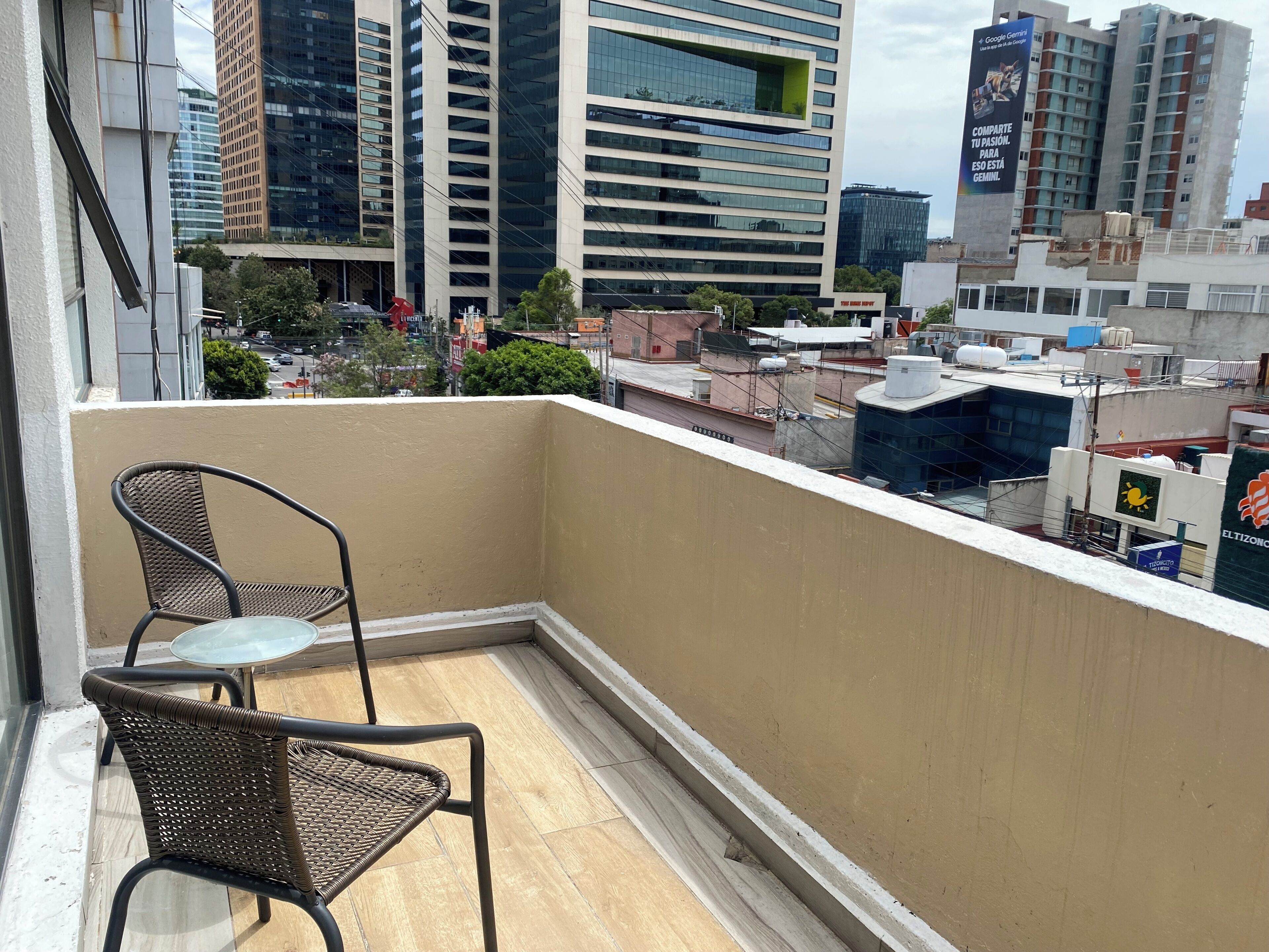 Een balkon met twee rieten stoelen en een glazen tafel, met uitzicht op een stadsbeeld met moderne gebouwen en een heldere lucht.
