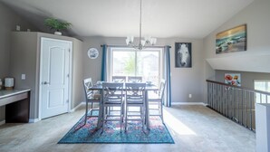 Un coin repas agrémenté d’un lustre, d’une table et de chaises. On y trouve une fenêtre avec des rideaux, une porte et un escalier.