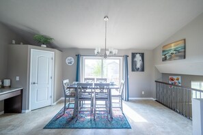 Een eetruimte met een kroonluchter, een eettafel en stoelen. Er is een raam met gordijnen, een deur en een trap.