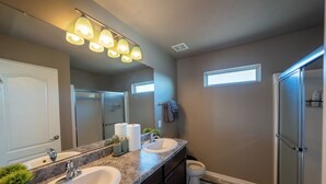 Une salle de bain avec deux lavabos, une douche et des toilettes. On y trouve des luminaires au plafond, un miroir et un porte-serviettes.