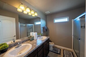 Een badkamer met twee wasbakken, een douche en een toilet. Er zijn inbouwspots, een spiegel en een handdoekhouder.