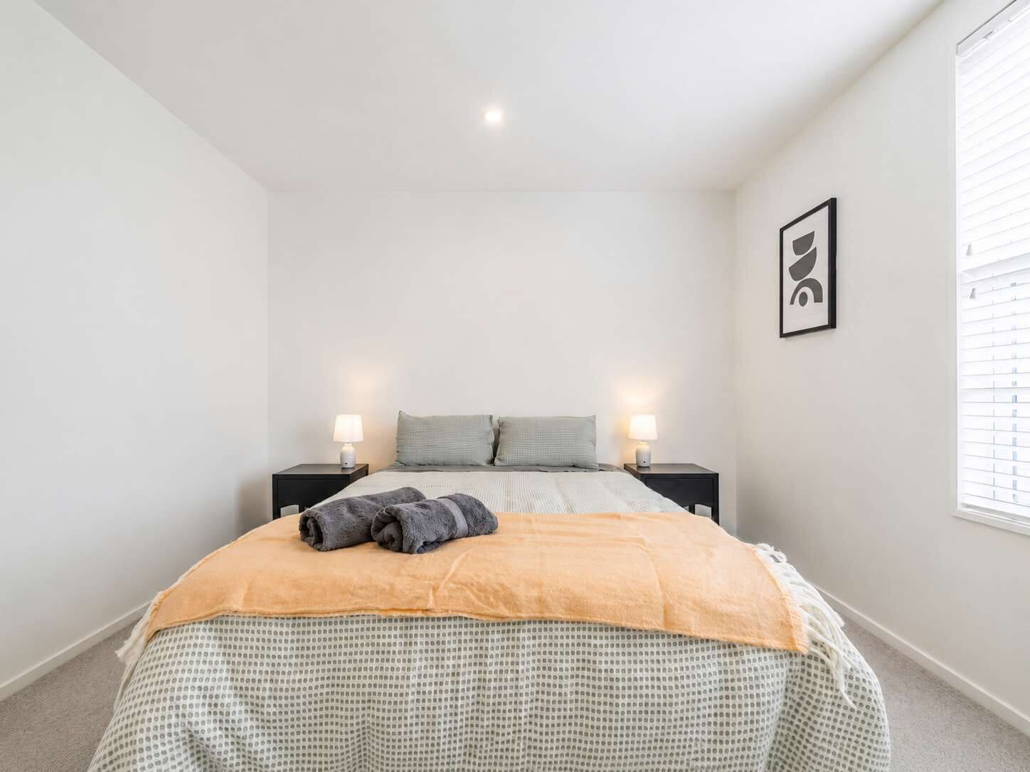 Ein Schlafzimmer mit einem Bett, zwei Nachttischen, einem Fenster mit Jalousien und einem gerahmten Bild an der Wand.