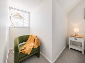 Ein Zimmer mit einem grünen Sessel, einer Stehlampe, einem Nachttisch mit Lampe und einem Fenster mit Jalousien.