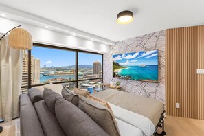 A modern hotel room with a large window offering a city view, a bed with a view, a sofa, and a wall-mounted TV. - 22F-Upscale & Modern-Financial District-Harbor View-1 Bedroom Condo w/Parking (Honolulu)