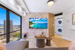A modern living room with a large TV, a sofa, a small coffee table, and a view of the cityscape. - 22F-Upscale & Modern-Financial District-Harbor View-1 Bedroom Condo w/Parking (Honolulu)