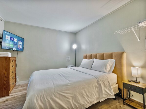 A hotel room with a bed, a wooden dresser, a TV mounted on the wall, a bedside lamp, and a wall-mounted drying rack.
