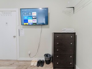 A room with a mounted television displaying various streaming services, a door with a coat rack, a dark-colored dresser, and a pair of slippers on the floor.