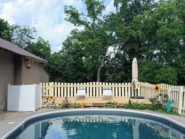 A swimming pool with a wooden deck, a covered patio, and a fenced area with outdoor furniture.