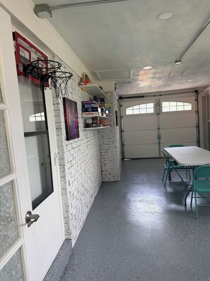A garage with a basketball hoop, shelves with board games, a table with chairs, and a door with frosted glass.