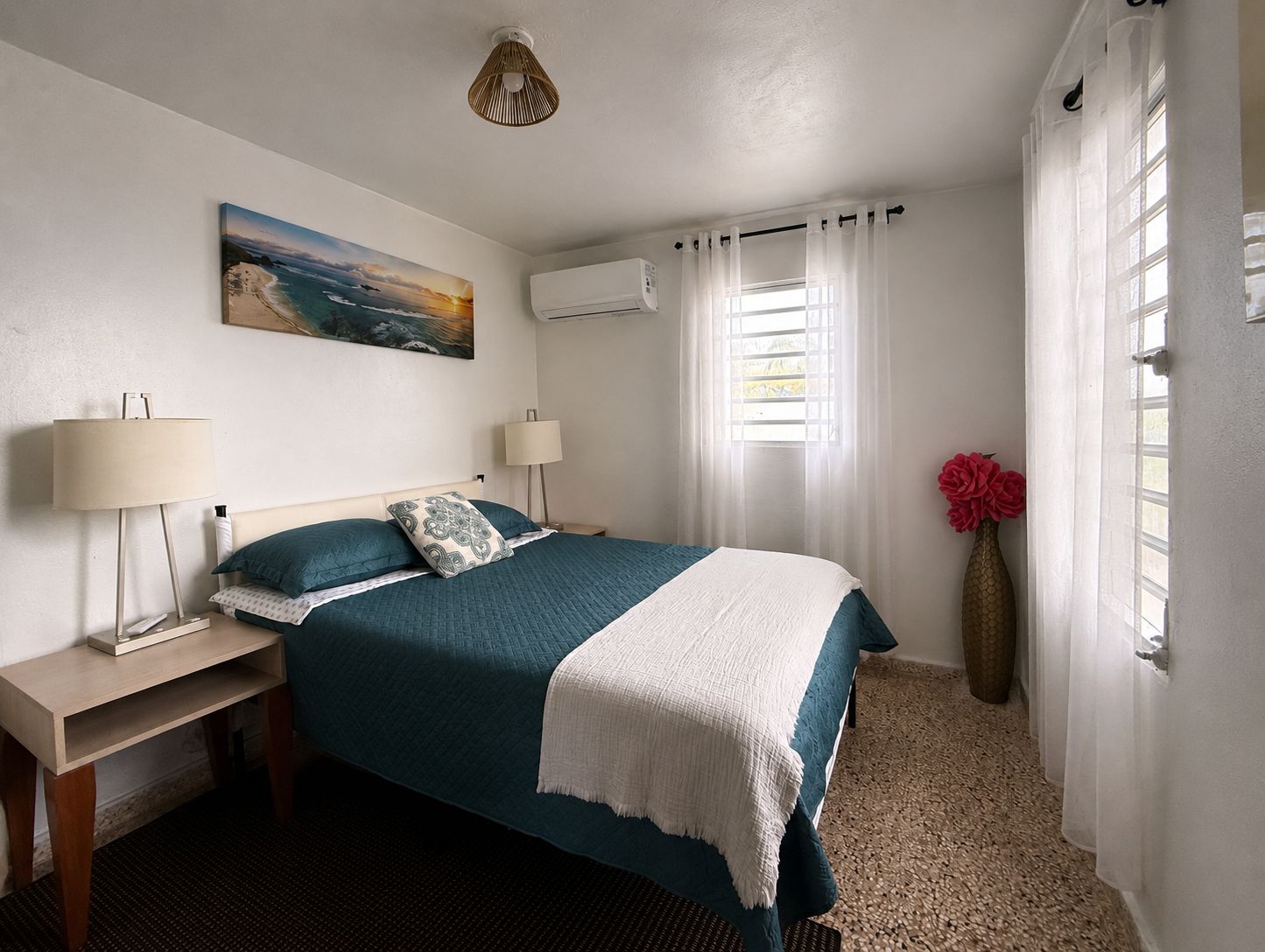 Ein Schlafzimmer mit Bett, Nachttischlampen, einem Gemälde, einer Klimaanlage, einem Fenster mit Vorhängen und einer Vase mit Blumen.