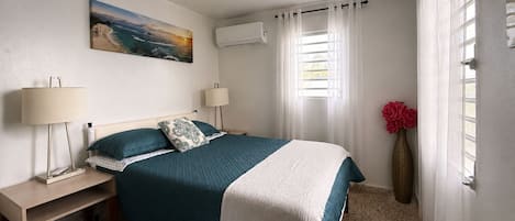 A bedroom with a bed, bedside lamps, a painting, an air conditioning unit, a window with curtains, and a vase with flowers.