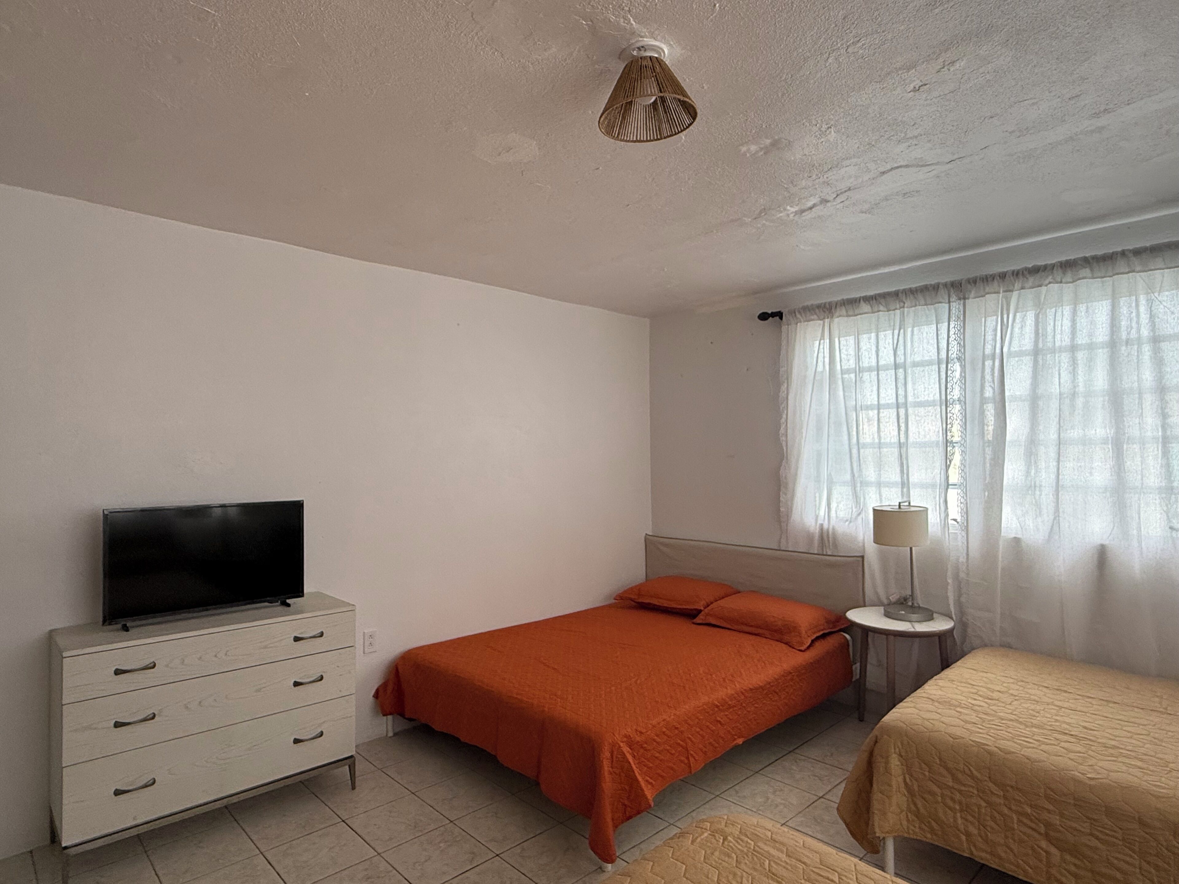 Un dormitorio con una cama, un televisor sobre una cómoda, una lámpara y un ventanal con cortinas.