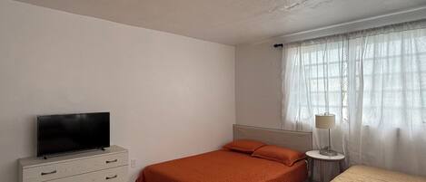 Un dormitorio con una cama, un televisor sobre una cómoda, una lámpara y un ventanal con cortinas.