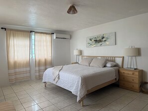 A bedroom with a bed, bedside tables, a lamp, curtains, a wall-mounted air conditioner, and a painting. - Caribe Social House-Stay • Celebrate • Connect (Loiza)