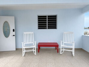 Una habitación con una puerta blanca, una ventana con persianas horizontales, dos mecedoras blancas y una mesa roja.
