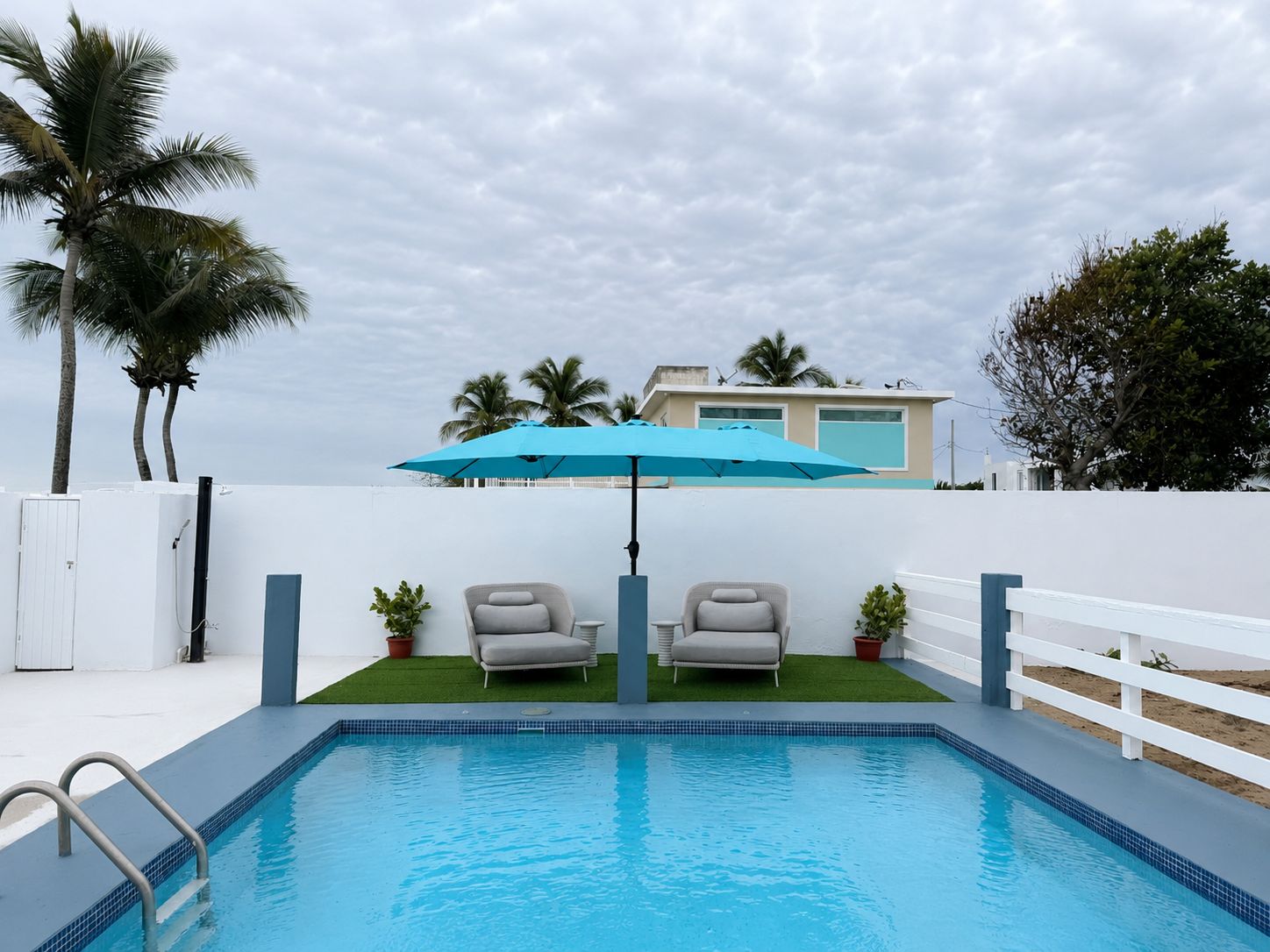 Ein Swimmingpool mit zwei Liegestühlen und einem blauen Sonnenschirm, umgeben von einem weißen Zaun und Palmen.