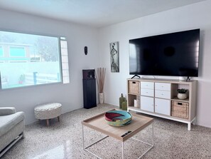Una sala de estar moderna con un televisor de pantalla plana grande, una mesa de centro con una cesta tejida, un sofá gris y un otoman pequeño y redondo.