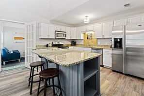 A modern kitchen with white cabinetry, a granite island, stainless steel appliances, and a blue sofa in the adjacent living area. - Miramar Home with Private Pool, Large Deeded Beach Access, Bikes & Paddleboards (Miramar Beach)