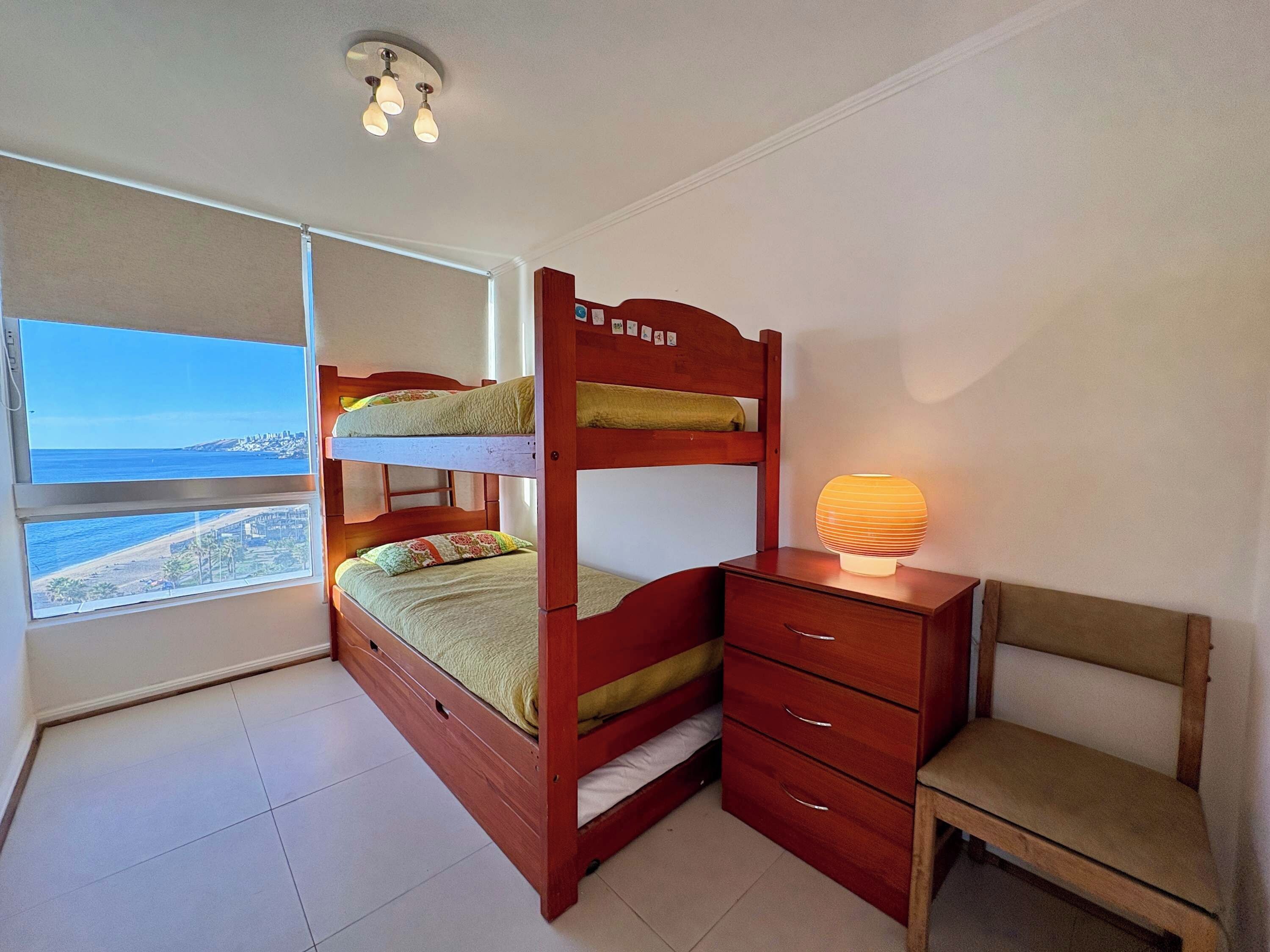 Una cama litera con escritorio y silla, una lámpara de noche y una ventana con vistas a la playa.