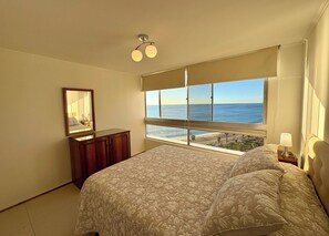 Ein Schlafzimmer mit einem großen Bett, einem Schminktisch, einem Spiegel, einem Nachttisch, einer Lampe und Meerblick durch ein großes Fenster.
